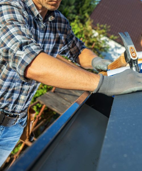 A carpenter diligently installs metal roofing on a residential building. The worker wears protective gloves and a plaid shirt, focusing on precision while using a hand tool.